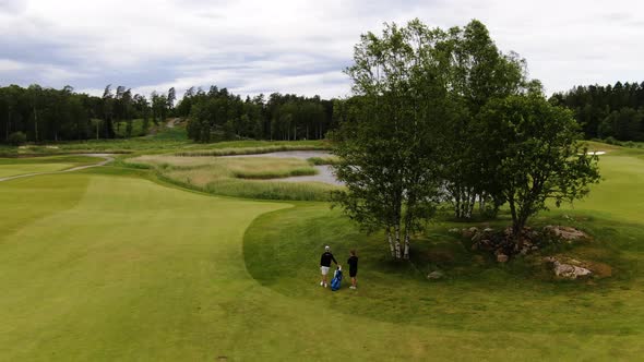 Golfer and caddie relaxing under tree of Swedish golf course. Aerial circling alt