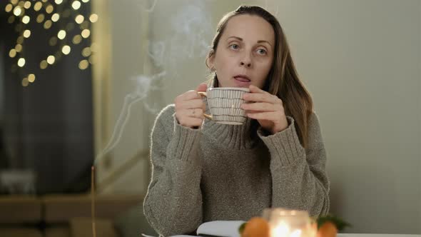 Young woman in a gray sweater drinks tea at home on a background of lights alt