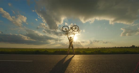 The Cyclist is Standing on the Edge of the Road and Lifting the Bike Above Him alt
