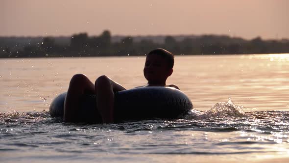 Silhouette of Happy Boy Swiming on Inflatable Circle in the River at Sunset alt