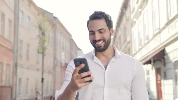 Smiling Man Using Mobile Phone And Walking Along City Street alt