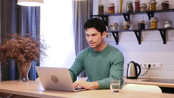 Man Celebrating Success While Working on Laptop in Kitchen alt