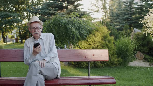 Mustachioed Senior Man Having Emotional Dispute on Web Cam on Bench in Park alt
