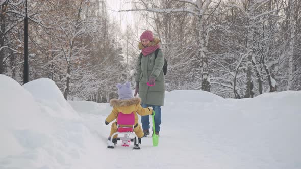 A happy mom pulls a snowcat sled with a baby in yellow warm clothes along a snowy path in the park. alt