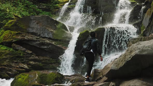 Girl Tourist Is Watching a Waterfall Standing High on Stones alt