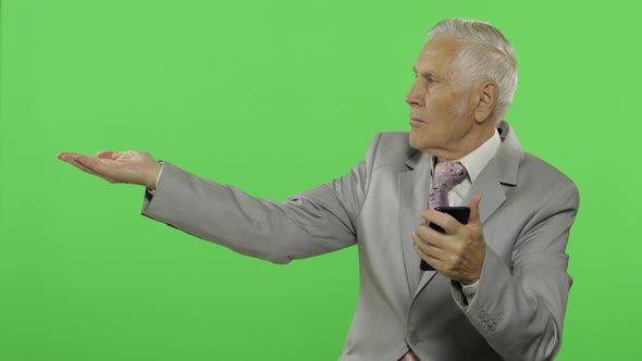 Handsome Elderly Businessman Texting on Smartphone and Points at Something alt