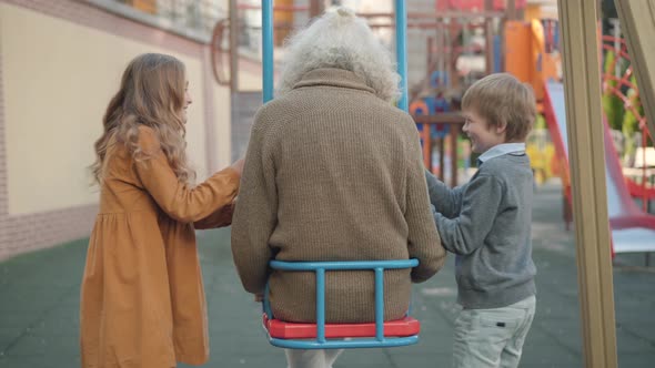 Back View of Relaxed Old Man with Long Grey Hair Sitting in Swings As Cheerful Little Boy and Girl alt