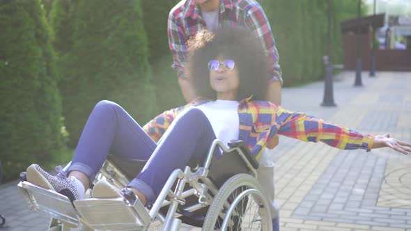 African Woman with an Afro Hairstyle Disabled in a Wheelchair in the Park for a Walk with a Friend alt