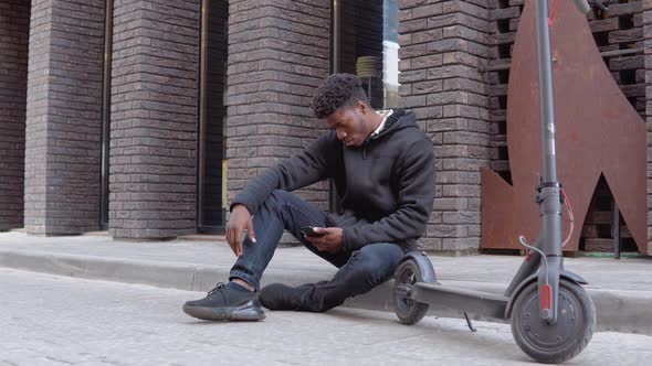 A Young African Man in a Black Sweater and Sneakers Sits on the Sidewalk Near a Building with a Dark alt