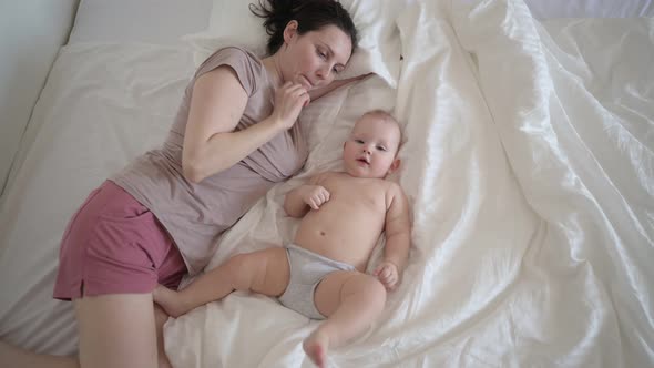Happy Young Mother Lying with Cute Infant Toddler Baby Boy on Bed Holding Him on Arms Hugging and