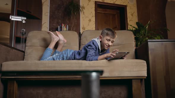 Smart Boy Lying on the Couch at Home and Watching Cartoon or Funny Video on Tablet PC alt
