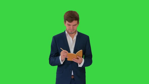 Young Businessman Thinking and Writing Notes in His Notebook While Walking on a Green Screen, Chroma alt