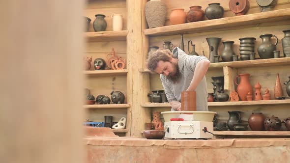 A young bearded potter works with a potter's wheel to create a tall brown jug alt