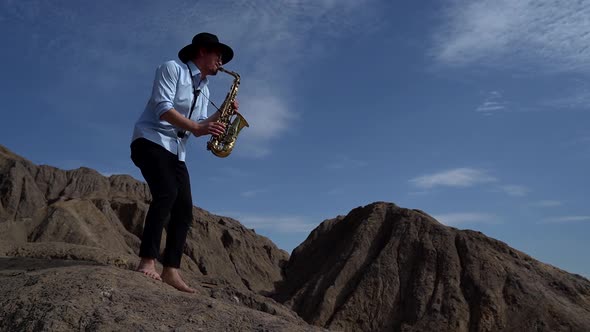 Barefoot Male Musician Plays a Wind Instrument Saxophone in the Mountains Against the Sky alt