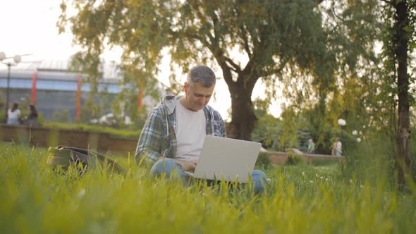 Man Works Behind a Laptop Sitting on the Grass At The Sunny Day
