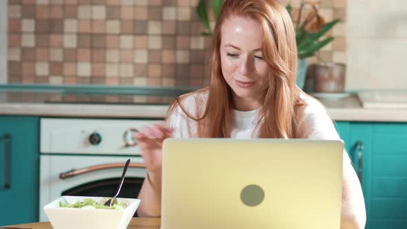 Cute woman with red hair works in the morning with a laptop in the kitchen. alt