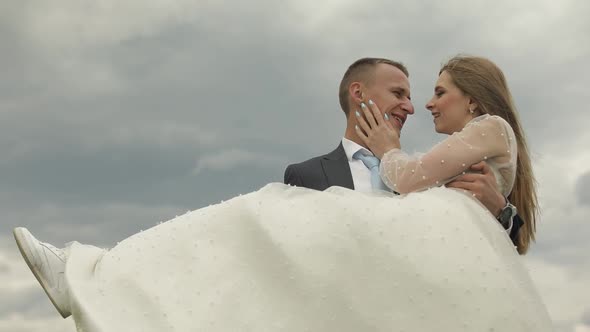 Groom Holds Bride in His Arms on Sky Background. Wedding Couple. Happy Family alt