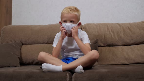 Sad Little Boy in a Medical Mask Sitting at Home on the Couch and Bored. alt