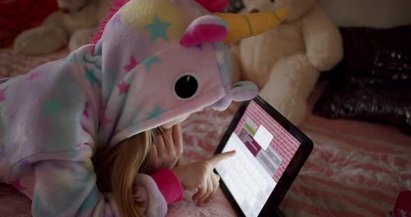 Little Girl is Playing in the Children's Room in Unicorn Costume Using Tablet alt