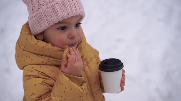 Child Girl Drinking Cocoa and Eating Cookie Outdoors in Winter Snowy Day alt