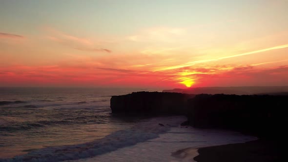 A red sky during sunset over the rocky cliffs while strong waves crash on the sandy beach near Londo alt