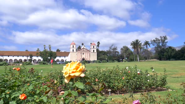 The Santa Barbara Mission building under blue skies with a foreground of flowers in the rose garden alt