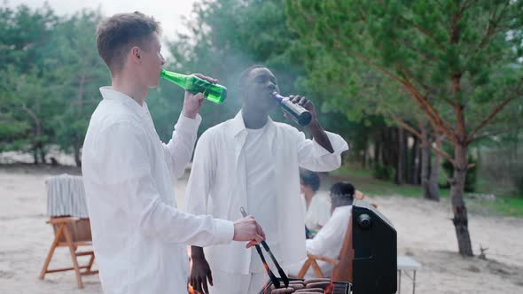 Handsome Young Men Cooking Barbecue at the Beach Party Drinking From Bottles alt
