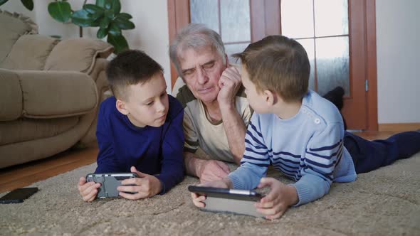 A grandfather with two children are lying and talking on the carpet in the middle of a room alt
