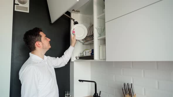 The caucasian man 30s puts dry, clean dishes in the cupboard alt
