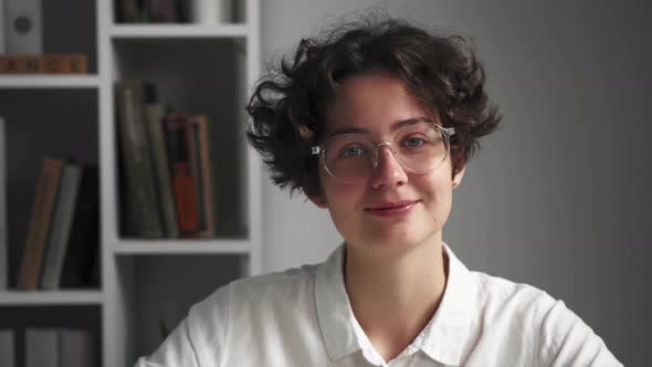 Portrait Of Smiling Dark Haired European Woman Standing In A Room Near A Bookcase. Happy Young Woman alt