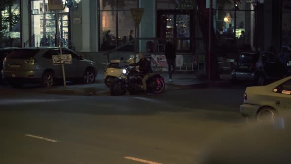 Attractive Rider Man Starting From Parking Place on Motorbike Turns on and Riding By Night City alt