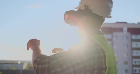 Engineer Builder on the Roof of the Building at Sunset Stands in VR Glasses and Moves His Hands alt