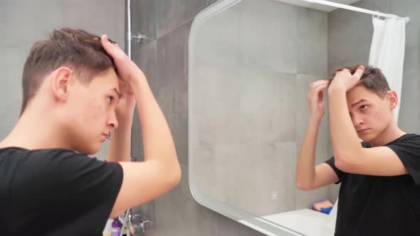 A Young Man in the Bathroom in Front of a Mirror is Styling His Hair alt