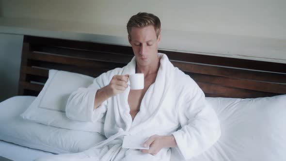 Young Man in Bathrobe Drinks Coffee in Bed Near Laptop alt