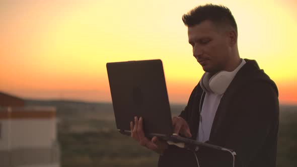 Standing at Sunset on the Roof with a Laptop and a Beer. A Man in a Hoodie Works Having Fun and alt