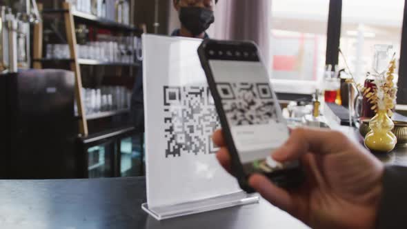 African american woman in face mask serving customer reading qr code with smartphone at cafe bar alt