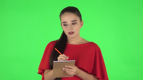 Portrait of Pretty Young Woman Is Standing and Thinking, Then Happy Writing with Pencil in Notebook alt