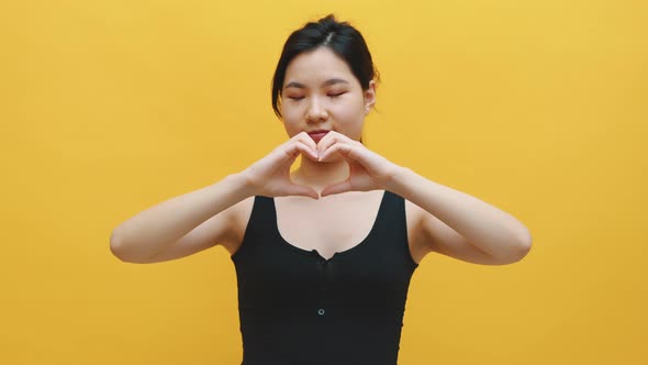 Happy Young Asian Woman Making Heart Shape with Her Hands. Isolated on Yellow Background alt