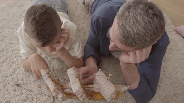 Portrait Cute Father and His Son Lying at Home on the Floor on the Fluffy Carpet Playing with Ship alt