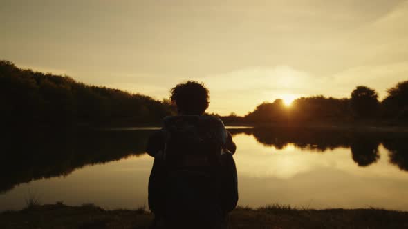 Boy with Backpack Sits Near the Lake at Sunset alt