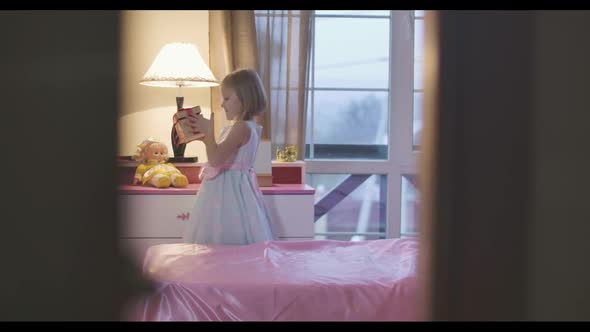 Shooting Through Doorway of Little Caucasian Girl Coming Up To Pink Bedside Table and Taking Casket alt