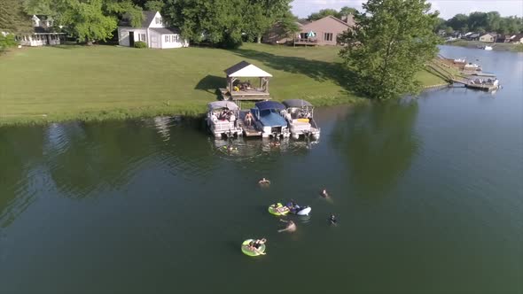 An orbiting aerial view of people swimming in a small lake. Pontoon boats docked on the shore. A man alt