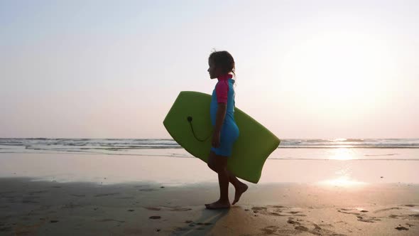 Little Girl Is Walking Along the Ocean Sand Beach with Surfboard at Sunset