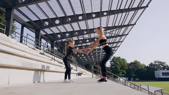 Two Attractive Sporty Girls-Elder and Younger Holding by Hands and Squating Outdoors Near Stadium alt