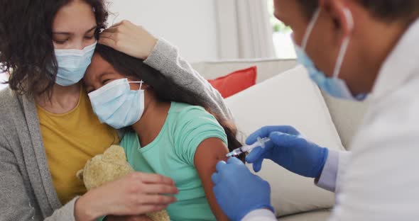 Hispanic male doctor making injection to girl with mother at home, all wearing face masks, alt