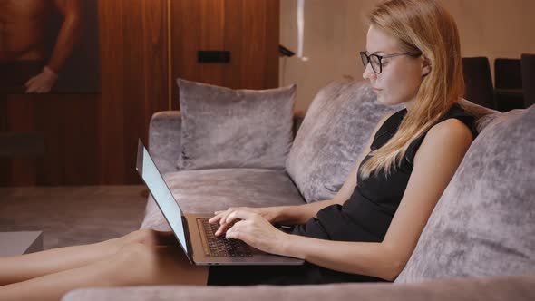 Woman in Evening Dress Sits on the Couch and Typing on Laptop on Her Lap alt