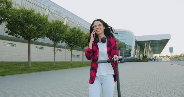 Woman in Casual Clothes which Walking with Her Personal E-scooter and Talking on Phone alt