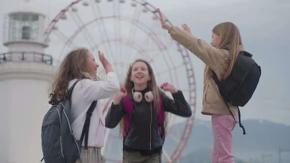 Peer Girls with Backpacks Jump Give High Five and Dance in Joy Near Giant Wheel