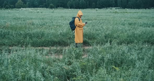 Man in a Yellow Raincoat Walks Through a Field and Looks at a Map in His Phone