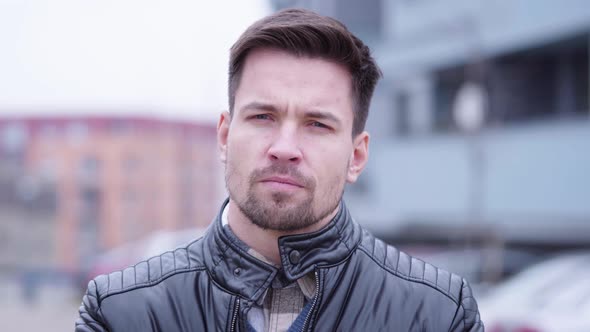 A Young Handsome Man Looks Seriously at the Camera in a Suburban Area with Buildings alt